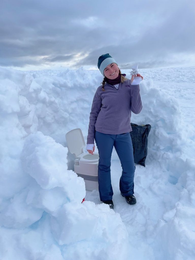 Camping in Antarctica. Girl holding toilet paper by toilet in the snow.